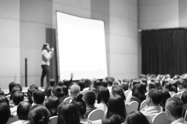 Seminar presenter at corporate conference giving speech. Woman speaker giving lecture to forum audience. Female executive manager leading discussion during technology company training event.