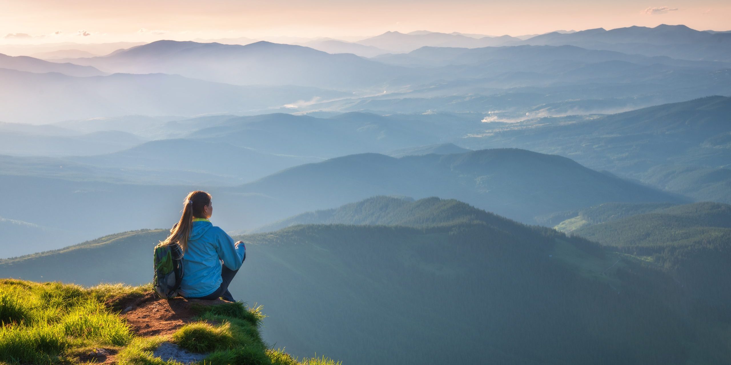 Young woman with backpack sitting on the mountain peak and beautiful mountains in fog at sunset in summer. Landscape with sporty girl, green grass, forest, hills , sky, sunbeams. Travel and tourism