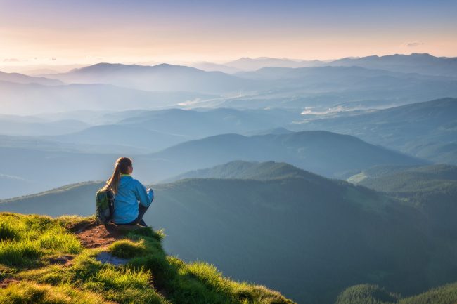 Young woman with backpack sitting on the mountain peak and beautiful mountains in fog at sunset in summer. Landscape with sporty girl, green grass, forest, hills , sky, sunbeams. Travel and tourism