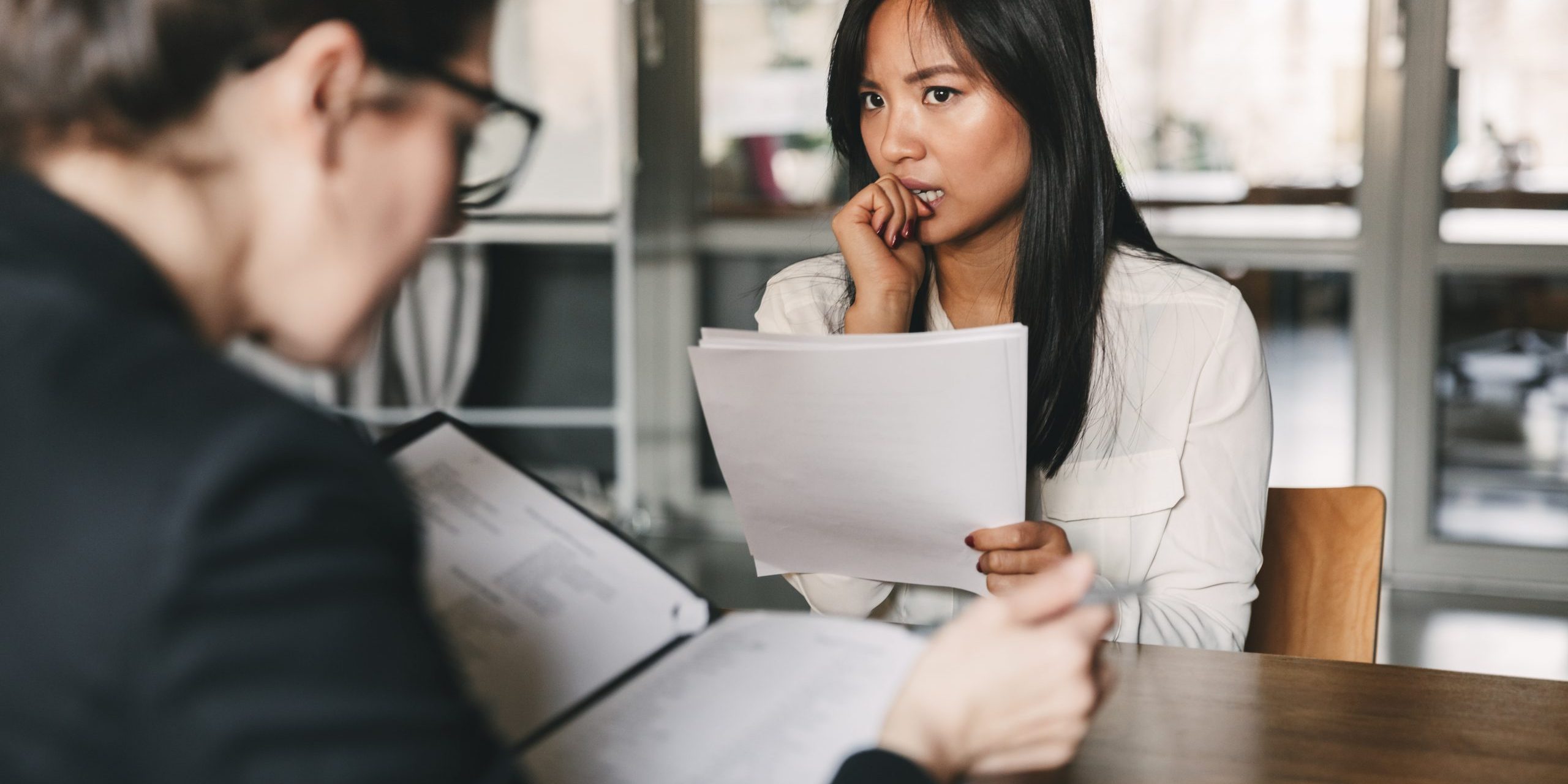 Image of concentrated tense asian woman looking at businesswoman, while sitting at table in office during job interview – business, career and recruitment concept