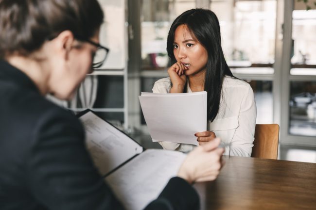 Image of concentrated tense asian woman looking at businesswoman, while sitting at table in office during job interview – business, career and recruitment concept