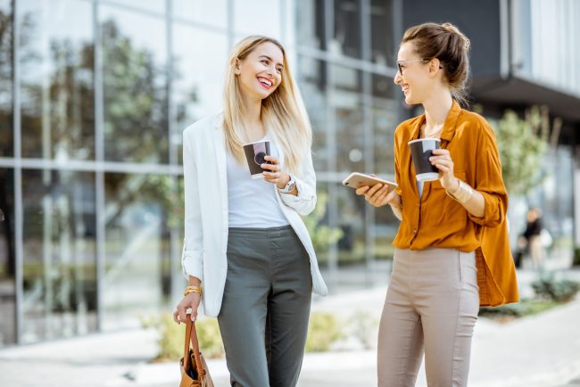 Two young businesswomen talking near the office building, having a small talk during the coffee break outdoors.